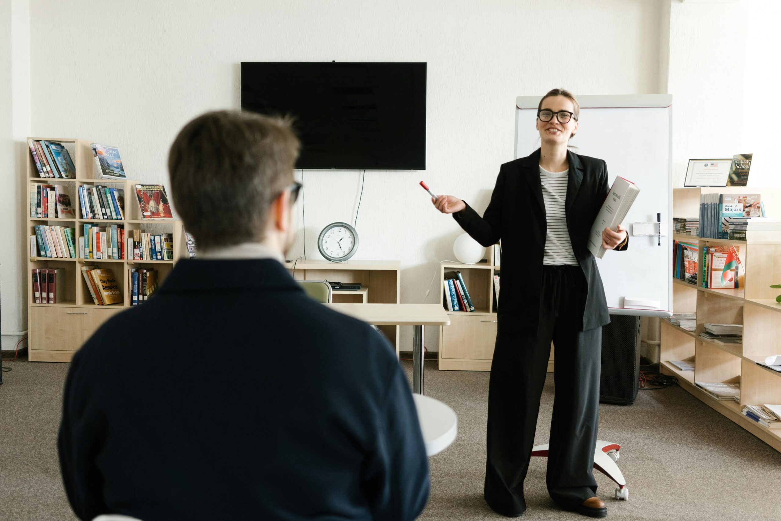 Home A teacher in a black blazer conducts a session with a student in a library setting.